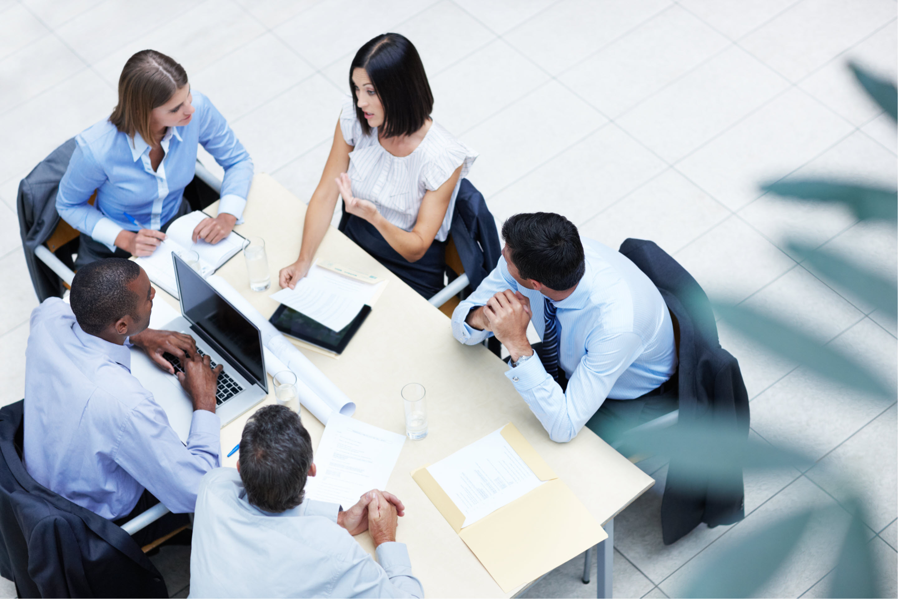 A group of business professionals with laptops, leaning in to a table mid-conversation. The table is shot from a birds-eye view, with a plant blurred in the periphery of the bottom right corner of the image.