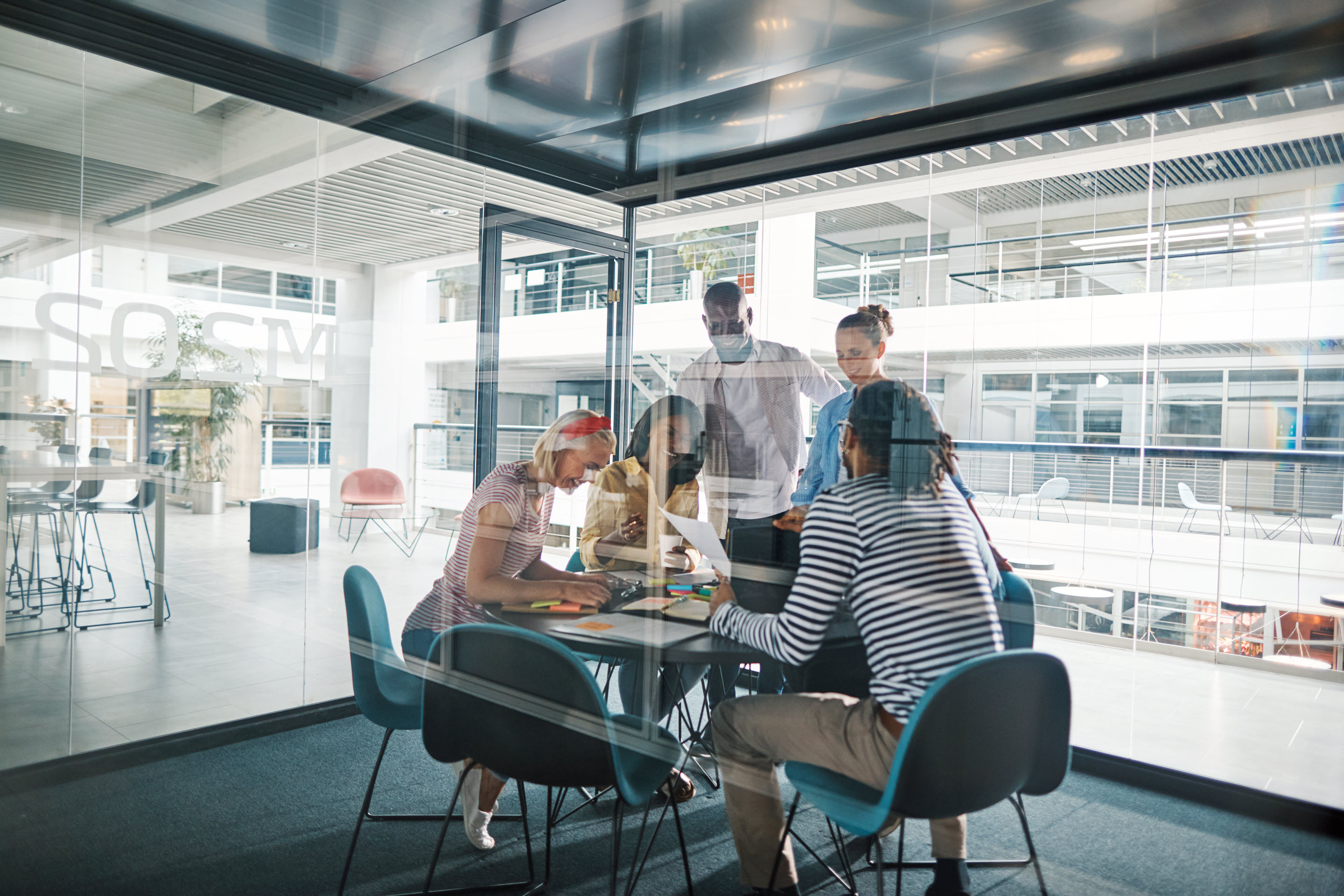 Through the glass wall of a meeting room, a group of business executives gather around a table to collaborate.