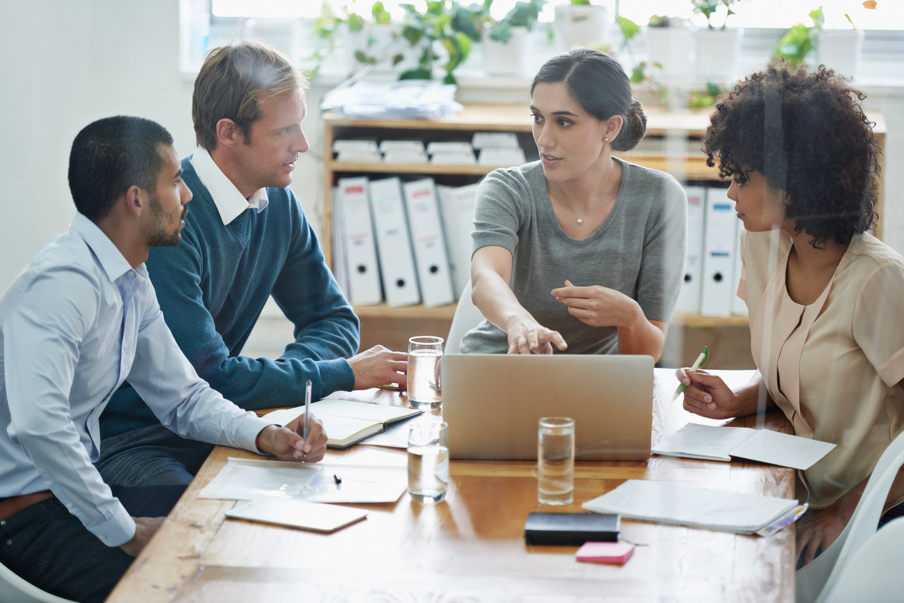 A group of business professionals gather around a table. The woman at the head of the table points to information on the tablet screen in front of her, turned to the others in the room, as if asking for collaboration or feedback.