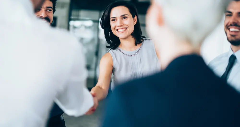 A businesswoman shakes the hand of another professional, making eye contact and smiling as she does so.