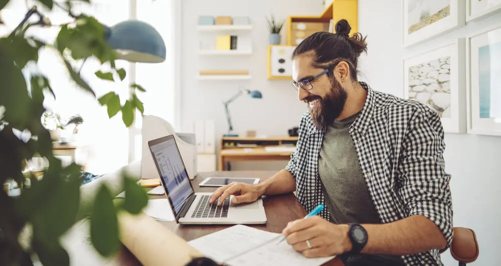 A man works from home, taking notes in front of his laptop. The office is entirely comprised of light colors, making it feel peaceful. A plant is visible but blurred in the foreground.