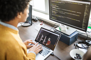 An over-the-shoulder view of a senior IT manager, dressed in yellow. They are working at their laptop, plugged into a monitor behind it, and looking at raw data for tax insights.