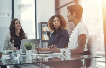 Three women executives sit at a conference room table, discussing business.