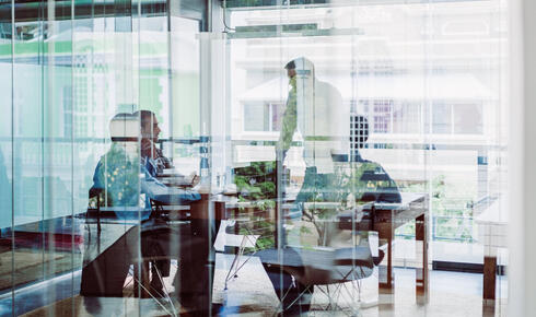 A group of business executives and thought leaders gather around a conference table for discussion. Part of the image is overlaid and obscured by the blue tone of reflection from the glass wall of the room.