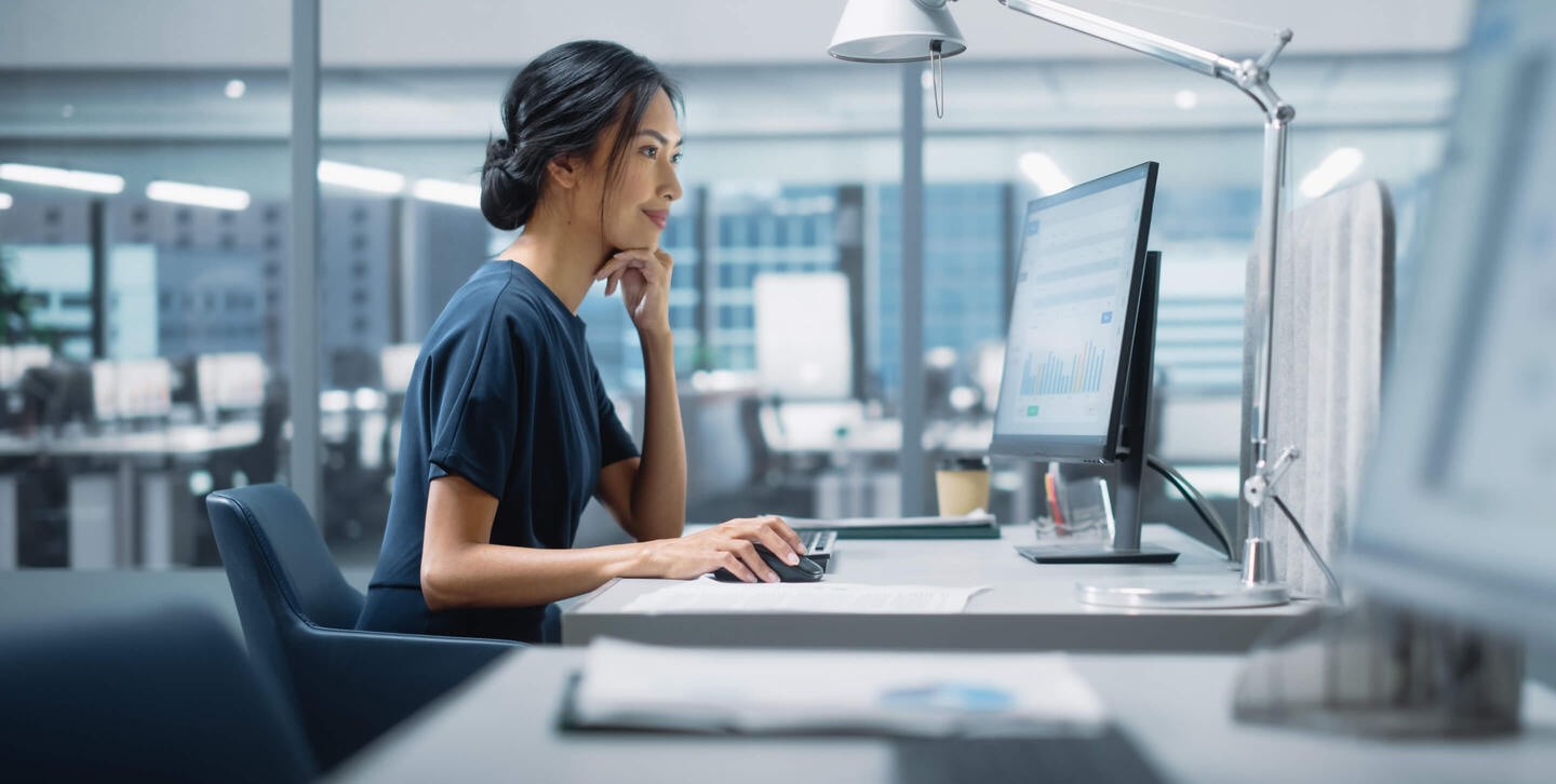 A side-view of a tax professional sitting at her desk, looking at analytics on her computer. The woman is leaned forward in a casual interest of the information on her screen. The image is primarily composed of the blues, grays, and whites of her open-floor office space, with a glass wall behind her separating her workspace from another work room filled with computers set up similarly to her workspace.