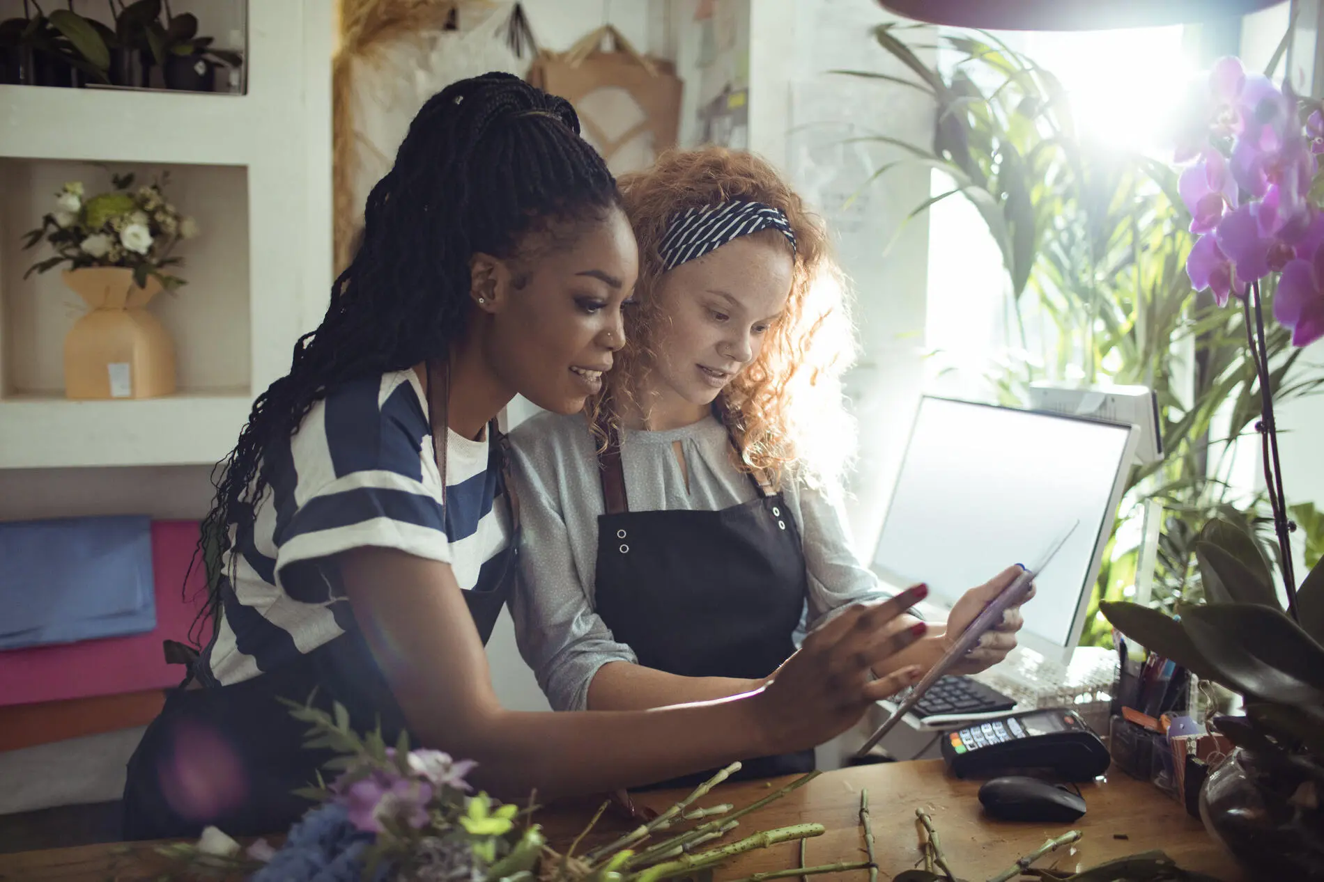 Two women in work aprons standing in front of their checkout computer in their flower shop. The woman in the foreground is showing her business partner information on a tablet. Light illuminates them from a window in the right side of the image, highlighting greenery and a string of purple flowers.